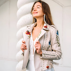 
A stylish luxury women's leather jacket featuring intricate floral embroidery, with a focus on the collar and sleeves. The model is smiling, wearing sunglasses, and showcasing the jacket's design and fit.