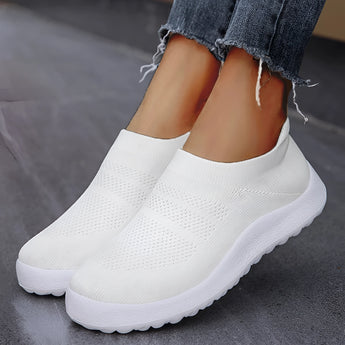 White women's slip-on sneakers with flexible sole, worn by a person; the sneakers are lightweight and breathable, showcased against a grey background.