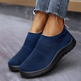 
A pair of blu slip-on sneakers for women, featuring a lightweight and breathable design with a flexible sole. The sneakers are worn by a person dressed in grey denim and sportswear, showcasing a casual fashion style.