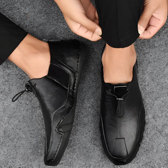 Men's beige suede slip-on loafers displayed on a model wearing black pants with crossed legs, highlighting the casual and breathable design of the shoes.