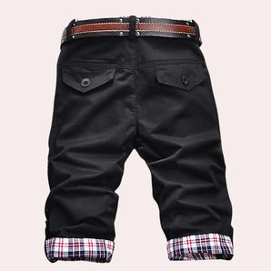 Men's black capri pants featuring plaid cuffs and a button pocket, worn by a model who pairs them with casual outerwear and a T-shirt for a stylish, sporty look.