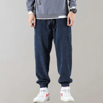 Men's black corduroy cargo pants with an ankle-tapered fit, styled urbanly, paired with a dark blue dress shirt.