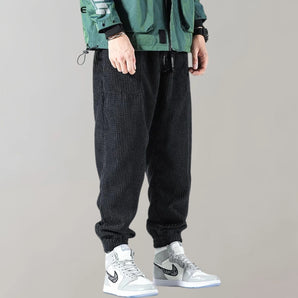 Men wearing black corduroy cargo pants with an ankle-tapered fit, styled in an urban fashion. The pants feature multiple pockets and are displayed in a standing pose.