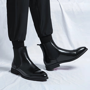 Men's black leather Chelsea boots, showcasing a formal slip-on design, worn on a human leg, emphasizing their ankle-height style and sleek appearance.