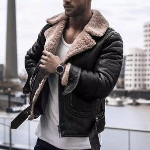 Men wearing a stylish black leather jacket with a shearling collar, paired with denim jeans. The man, who has a beard and is wearing eyewear, is standing outdoors in front of a building. The jacket is urban fashion-themed, and the image is captured using flash photography.