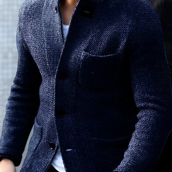 Men's charcoal tweed blazer in a slim fit design, featuring a classic herringbone pattern, modeled by an individual wearing a marine blue dress shirt underneath. The blazer has a structured collar, long sleeves, and is complemented by a watch on the wrist. The overall attire exudes a sophisticated and formal look.