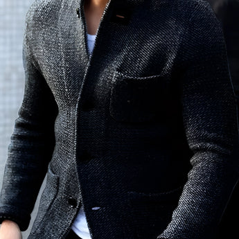 Men's charcoal tweed blazer in a slim fit, featuring a herringbone pattern. The jacket has a classic collar, long sleeves, and is shown in a Nero color in size 4XL. The outerwear is paired with a dress shirt, and the model has a beard and is wearing a watch.