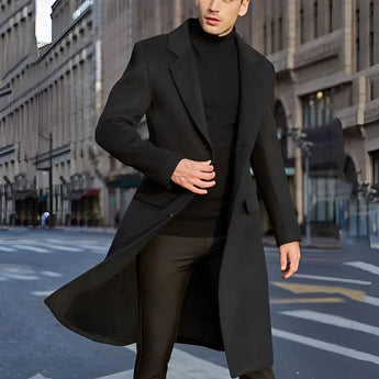 A man wearing a stylish urban fashion overcoat in grey wool, standing confidently. The coat features long sleeves and a structured shoulder design, epitomizing winter fashion. The individual is accessorized with sunglasses, enhancing the modern and sophisticated look.