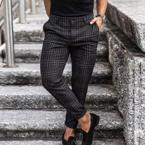 Men's slim-fit dark gray plaid trousers, ankle-length, styled with black shoes, a white shirt, and a black outerwear jacket.