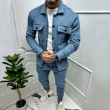
A man standing indoors wearing a two-tone blue lightweight tracksuit, featuring long sleeves and a collar, in a stylish urban setting. He is possibly in a casual loungewear context, with a houseplant visible in the background. The tracksuit is labeled as "Azzurro" and is sized 3XL.