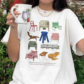 White Novelty Chair Graphic Tee in 2XL featuring a quirky furniture design, worn by a smiling person making a gesture with their hand, with plant elements in the background.