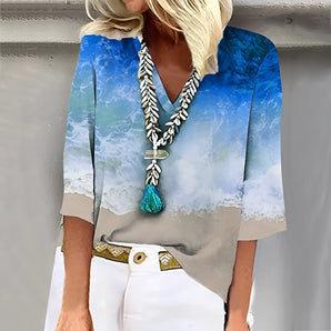 A plus-size blue v-neck blouse with a beach scene design, featuring a tassel necklace, displayed on a hanger against a white background.
