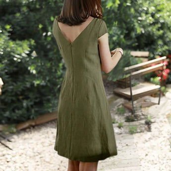Olive green linen knee-length A-line dress with a V-neck and short sleeves, featuring a model standing outdoors near a bench and grass.