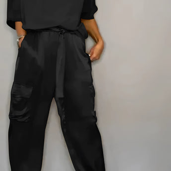 Olive green satin jumpsuit with a luxurious, smooth finish, featuring a relaxed fit suitable for designer loungewear. The jumpsuit has long sleeves, a tailored waist, and a V-neck, shown here in a Nero color and 5XL size.