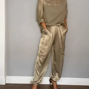 Olive green satin jumpsuit featuring long sleeves, a cinched waist, and a luxurious design. The jumpsuit is worn by a model with a smiling expression, showcasing the elegant fit around the thighs and legs, highlighting its status as designer loungewear.