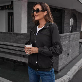 Olive green women's cropped utility jacket worn by a model sitting on an outdoor bench, paired with jeans and sunglasses, showcasing a lightweight street style look.