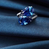 A woman's hand wearing an ORSA 925 Sterling Silver Double Teardrop Blue Topaz Ring, showcasing its shimmering azure gemstones set in a silver band, captured in flash photography.