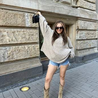 Oversized beige chunky knit turtleneck sweater worn by a model, showcasing cozy winter fashion. The model is accessorized with sunglasses, highlighting a stylish casual look.