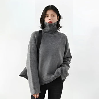 
A woman wearing a dark gray oversized turtleneck sweater with chunky knit texture, featuring a high collar and long sleeves, suitable for winter. The sweater is styled for a relaxed fit, emphasizing casual comfort and warmth.