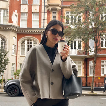 Oversized gray blazer paired with a cropped top and trousers, styled as a chic streetwear outfit. The model is wearing sunglasses and standing near a window, showcasing a sophisticated urban look.