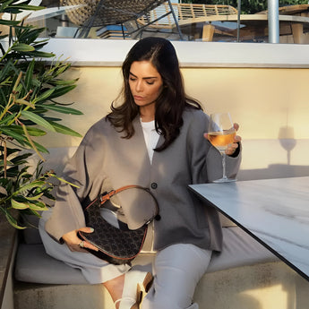 Oversized gray blazer paired with a cropped top and trousers, worn by a seated model. The outfit exudes chic streetwear style, complemented by fashionable eyewear. The setting includes a minimalist backdrop with a touch of greenery.