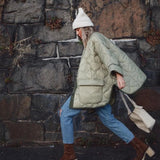 Woman wearing an oversized green quilted puffer jacket, lightweight and suitable for casual autumn wear, standing on a road surface, paired with jeans and sneakers, holding a bag.