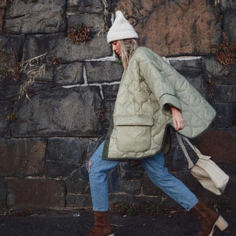 Woman wearing an oversized green quilted puffer jacket, lightweight and suitable for casual autumn wear, standing on a road surface, paired with jeans and sneakers, holding a bag.