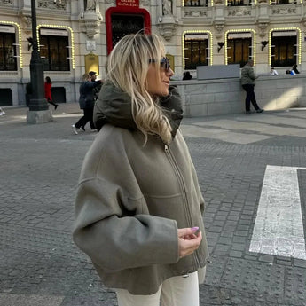 Oversized khaki hooded jacket worn by a woman standing against an urban backdrop with windows and buildings, showcasing minimalist fashion with long sleeves.