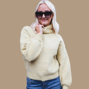 Oversized pale yellow chunky knit turtleneck sweater modeled by a smiling person wearing glasses, with arms raised to show the long sleeves, capturing a cozy fall fashion look.