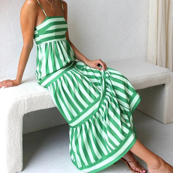 Pink and blue striped spaghetti strap maxi dress designed for summer beachwear, showcasing a flowing silhouette with a fitted waist and ankle-length hemline. Model accessorizes with casual sandals.
