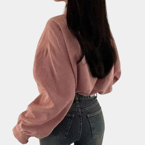 A woman wearing a pink cropped V-neck blouse with puffed sleeves, paired with jeans. She is accessorized with sunglasses and is making a gesture with her arm. The blouse features a stylish collar, complementing the casual chic look.