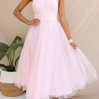 Pink halter neck backless tulle dress showcased in a wedding or summer event setting, featuring a flowing design with delicate shoulder straps. The dress is surrounded by a backdrop of greenery and flowerpots, highlighting its elegant and airy style.