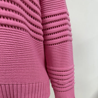 
A pink textured ribbed sweater with a cut-out design, featuring long sleeves, shown on a person. The sweater has a modern knitwear style and is crafted from woolen material.