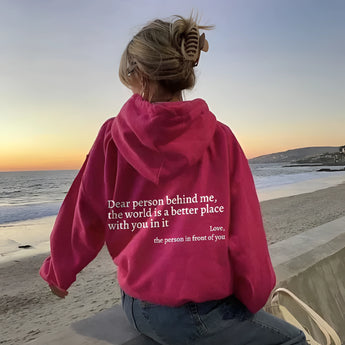 Pink tie-dye hoodie with the words "Self-Love" printed on it, worn by a smiling individual standing on a beach, with a clear sky and ocean in the background, conveying a sense of happiness and leisure.
