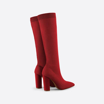 Red knee-high sock boots with high heels made from ribbed fabric, shown on a model's legs seated in a chair.