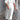 
A woman wearing a white one-piece maxi dress with puff sleeves and a square neck, featuring studded pockets, standing and smiling.
