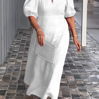 
A woman wearing a white one-piece maxi dress with puff sleeves and a square neck, featuring studded pockets, standing and smiling.