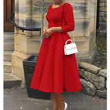 Woman wearing a red midi dress with long sleeves, a boat neck, A-line silhouette, and pockets, standing and showcasing the fit around the waist, size 2XL.