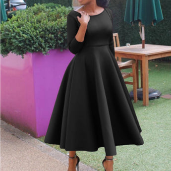A woman wearing a red midi dress with long sleeves, a boat neck, and an A-line silhouette, standing and showcasing the dress's pockets.