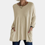 
A woman wearing a khaki oversized tunic sweater with pockets, suitable for fall fashion. The sweater features a relaxed fit with long sleeves, a round neckline, and covers the hips. The color appears to have a grey undertone.