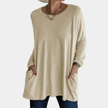 
A woman wearing a khaki oversized tunic sweater with pockets, suitable for fall fashion. The sweater features a relaxed fit with long sleeves, a round neckline, and covers the hips. The color appears to have a grey undertone.