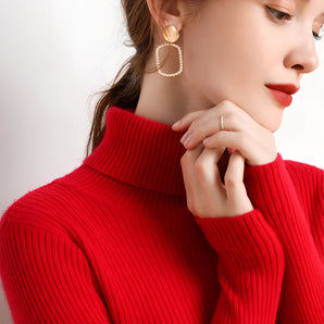 
A woman wearing a red ribbed turtleneck sweater in size 2XL, accessorized with gold pearl hoop earrings. Her hair is styled neatly, and she has makeup on, including lipstick and eye makeup.