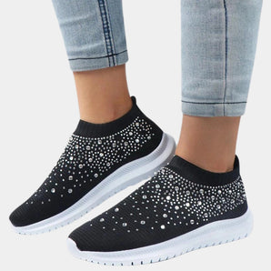 
A pair of women's fashion slip-on sneakers featuring a black mesh design adorned with rhinestones, displayed on a person's feet. The sneakers are styled with casual grey sportswear and denim, highlighting their versatility and trendy appeal.