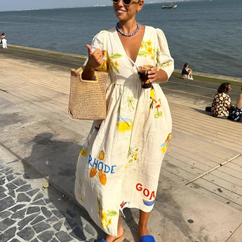 Woman wearing a beige RHODE dress with a fruit pattern, standing outdoors under a blue sky, holding a bag and wearing sunglasses.