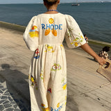 Woman wearing a RHODE dress, a women's cotton printed summer maxi with a fruit pattern, standing outdoors under a clear sky.