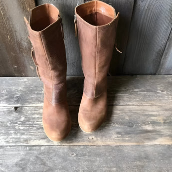 Rustic women's distressed brown leather mid-calf boots with a zipper, shown on a human leg, styled against a wooden backdrop with a flowerpot in the background.