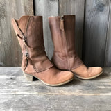 Brown distressed leather mid-calf boots with zippers, featuring a rustic design, styled as cowboy or riding boots, shown on a wooden surface.