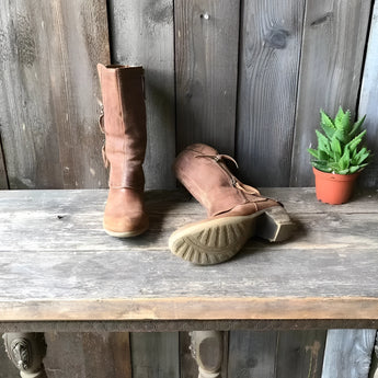 Rustic women's distressed brown leather zipper mid-calf boots placed on a wooden floor next to a potted houseplant.