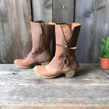 Rustic women's distressed brown leather mid-calf boots with a zipper, set against a neutral background with subtle wood and plant elements.