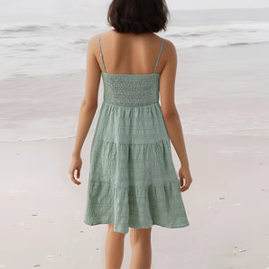Sea green smocked backless tiered women's summer dress worn by a woman near water, with visible shoulders and arms, highlighting the dress's flowing one-piece design.